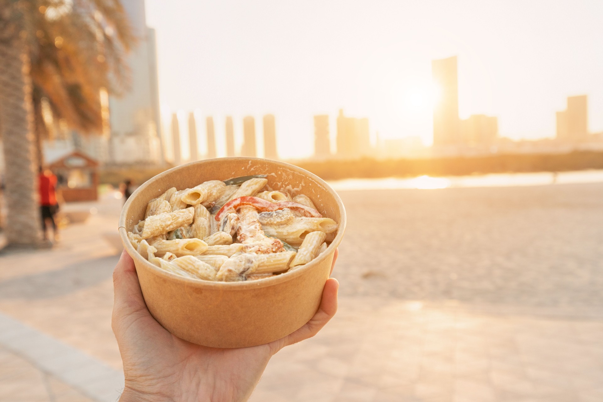 Hand holding a bowl of delicious penne pasta with vegetables in Abu Dhabi, United Arab Emirates, with the city skyline in the background at sunset